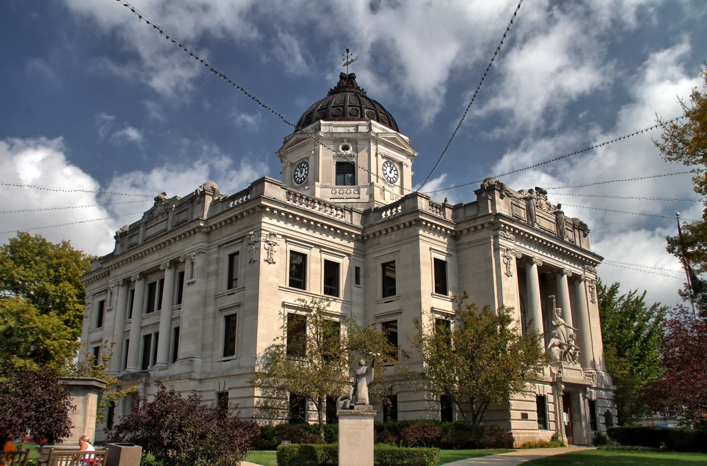 Exterior view of the Monroe County Courthouse in Bloomington, Indiana—a Beaux Arts limestone building with a grand colonnaded entrance, a central dome topped by four clock faces and a fish‑shaped weather vane, framed by trees under a partly cloudy sky.